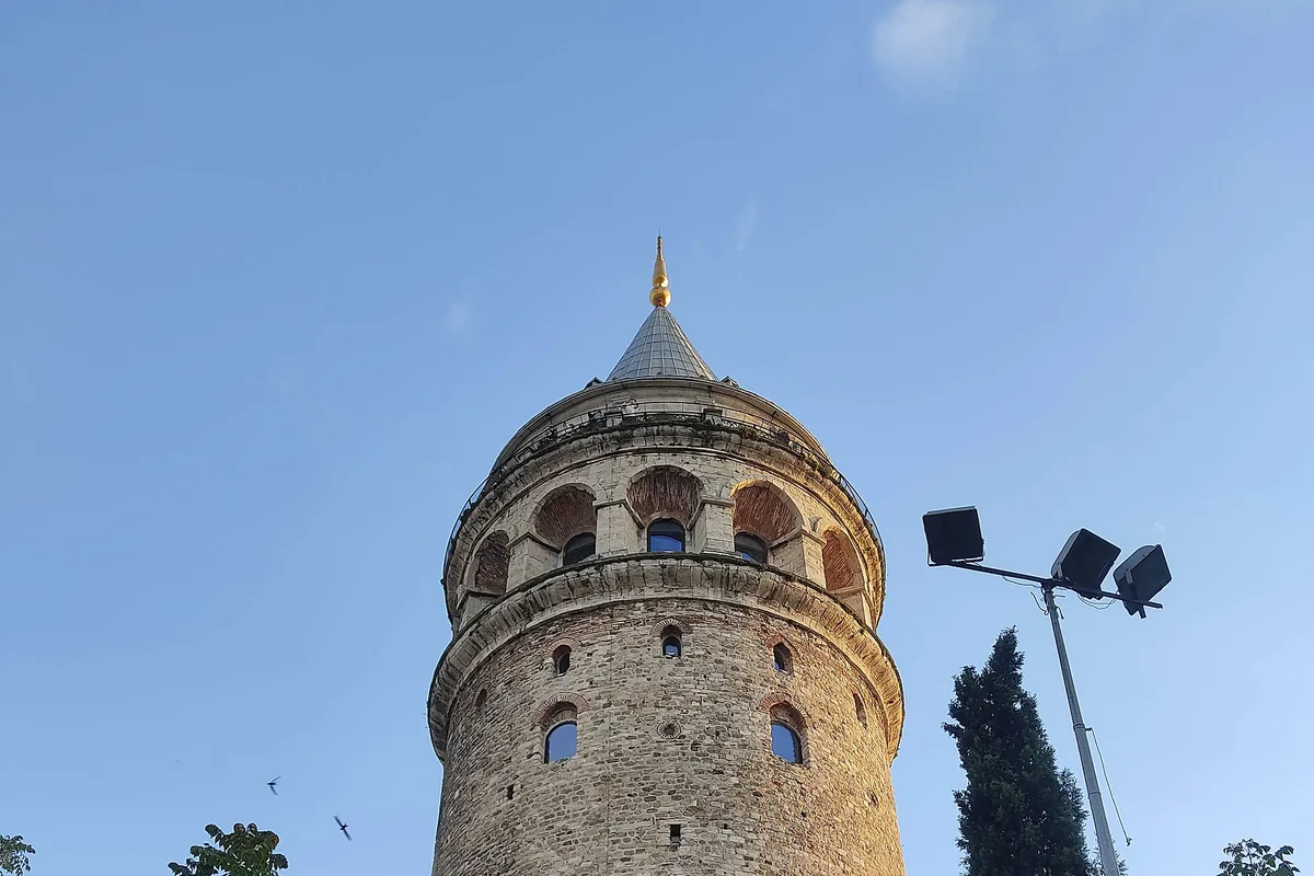 The stone drum and cone roof of Galata Tower against a blue sky, with a lamp post at the right.