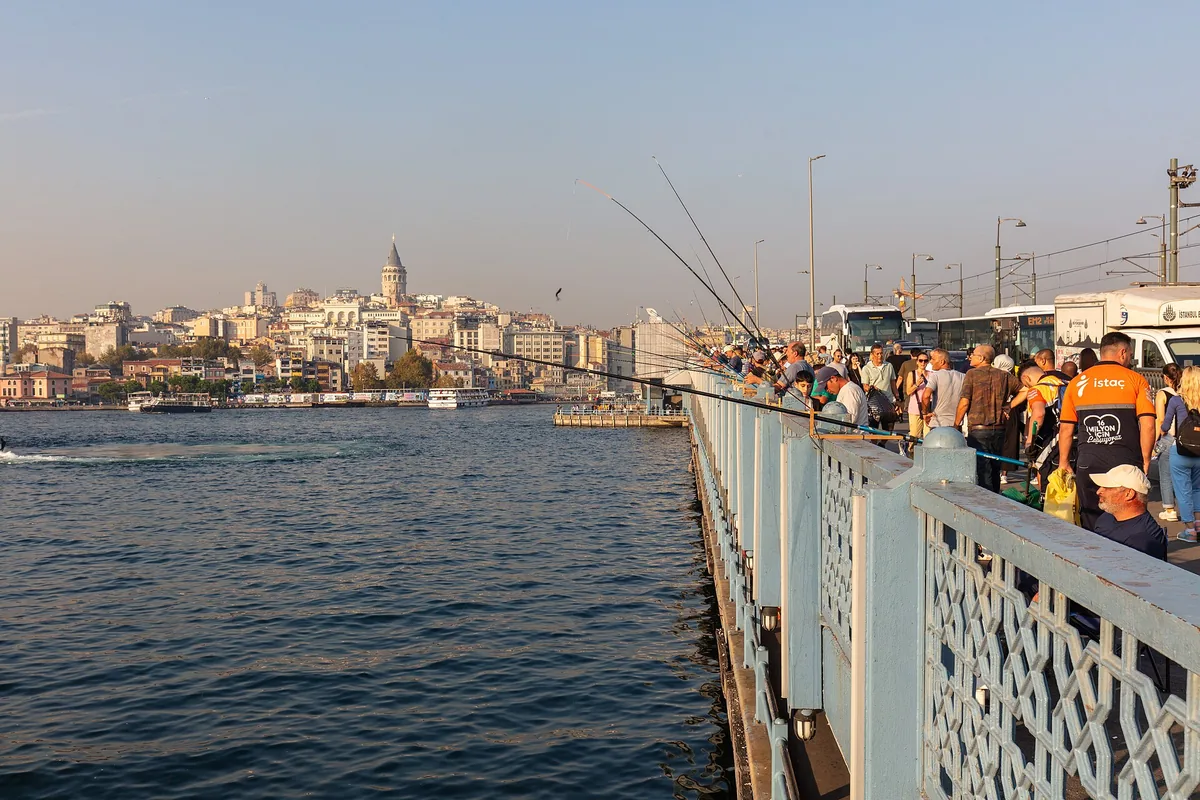 Anglers along the railing of the Galata Bridge, with the Galata hill and its tower in the distance across the water.