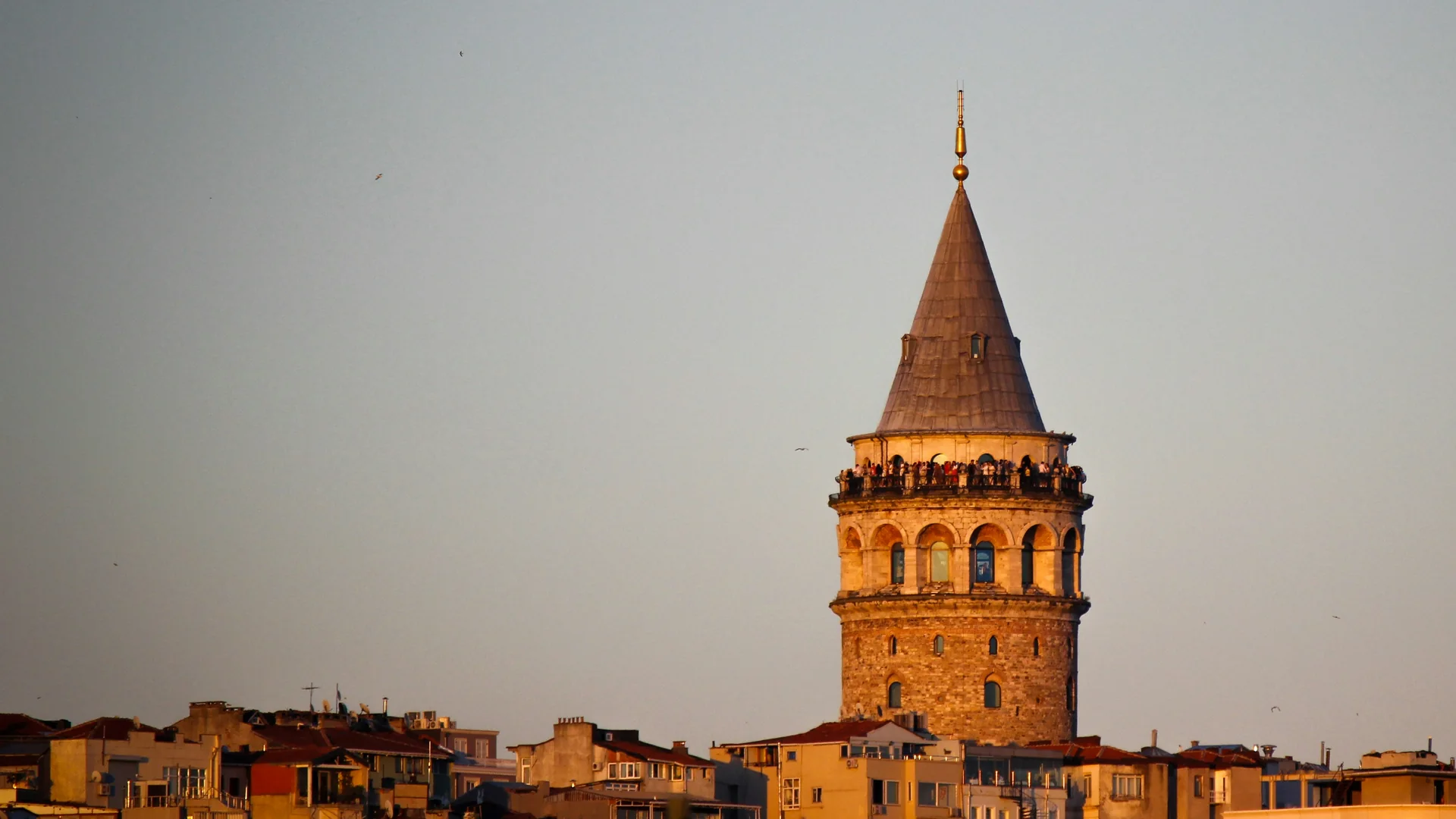 Galata Tower at golden hour, its cone roof and stone drum rising above Beyoğlu rooftops, with visitors along the balcony beneath the cone.