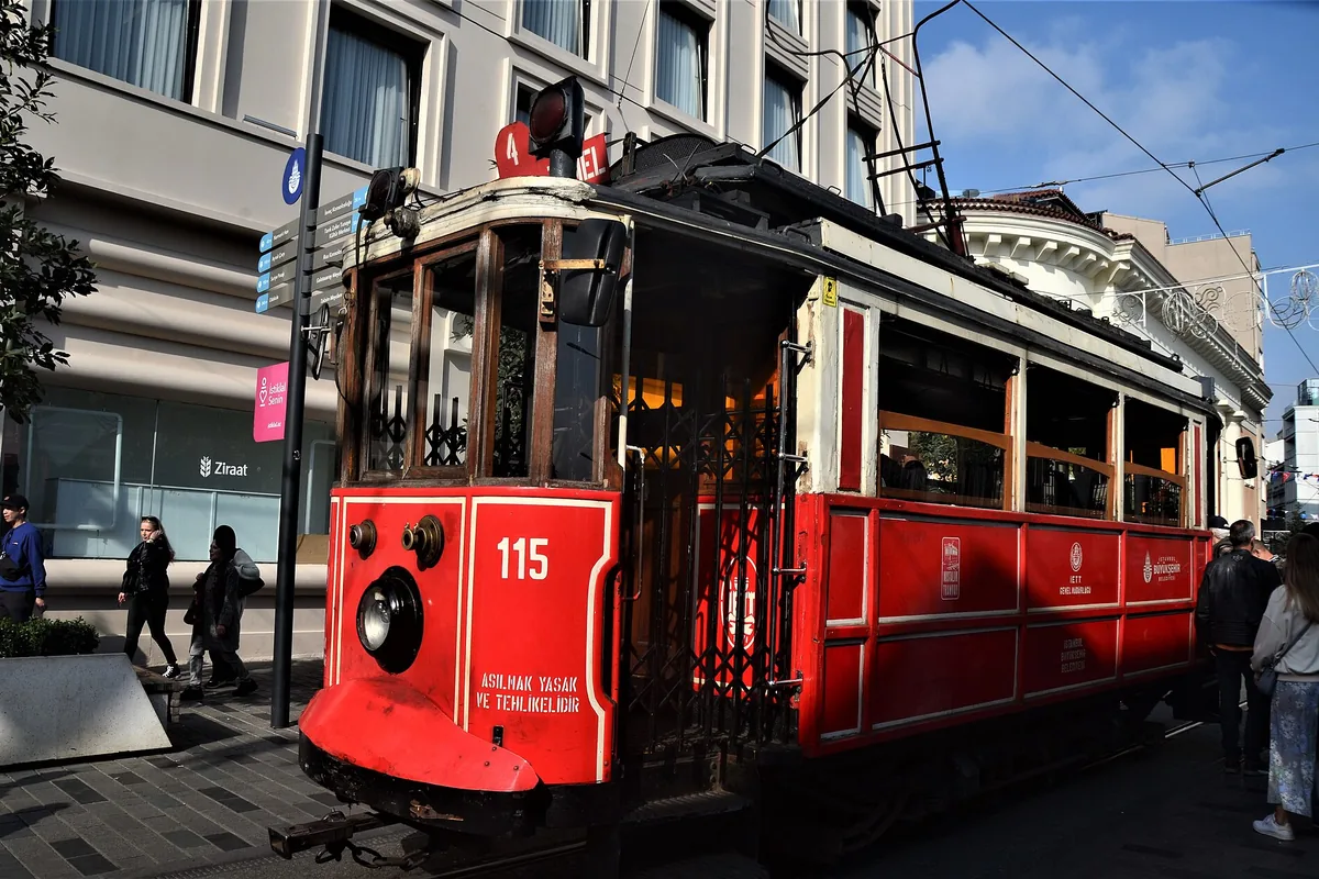 A red nostalgic tram on İstiklal Avenue with overhead wires and a pale apartment block behind it.